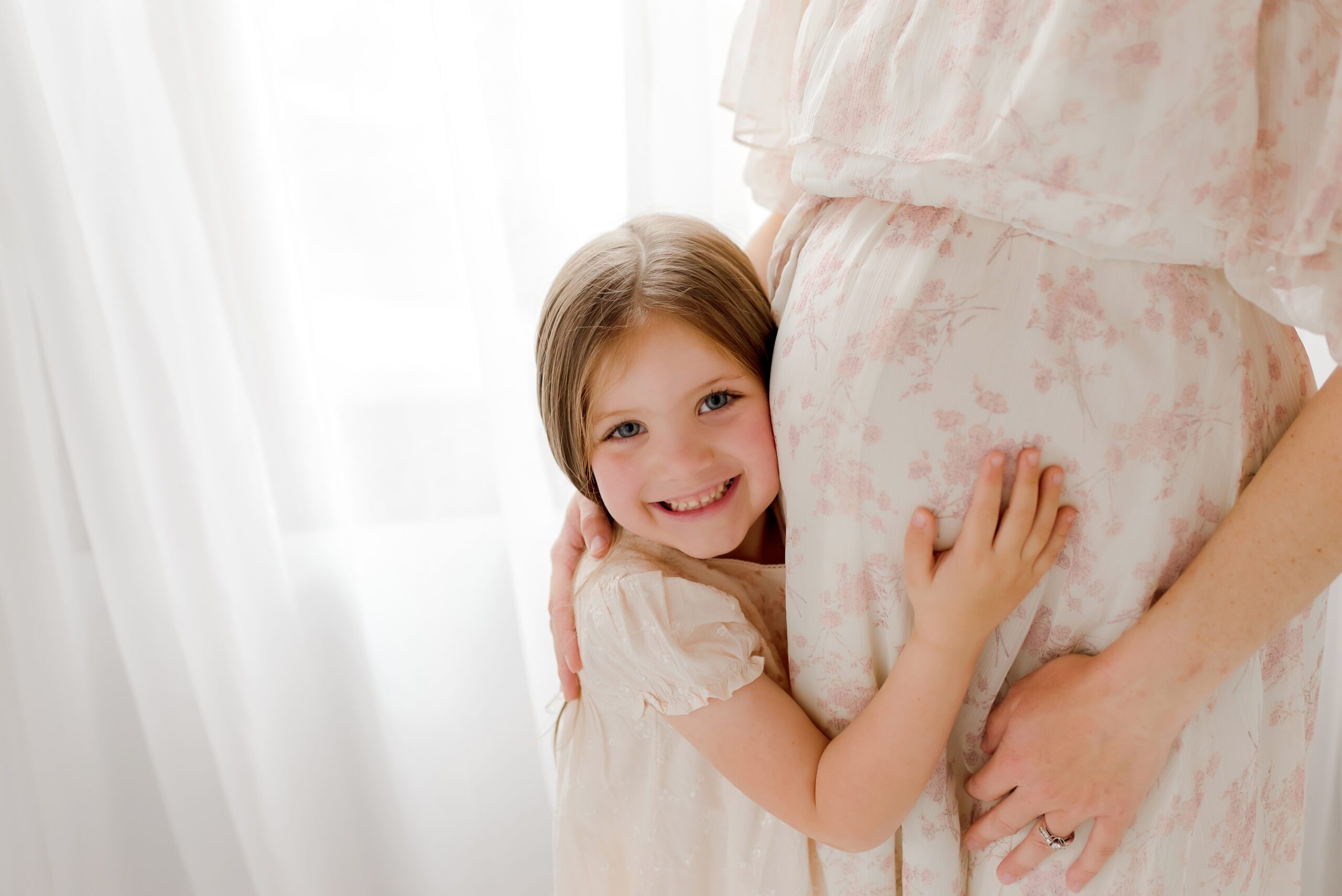Young girl smiling as she hugs her mother's pregnant belly, posed in front of a large window with sheer white curtains.