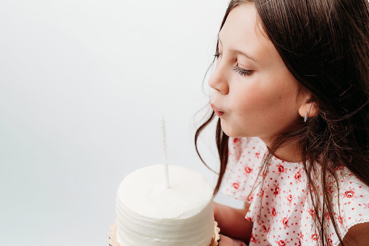 Girl in pink floral dress blowing out a birthday candle on a white cake during birthday session at Kelli Dease Photography, Farmington CT