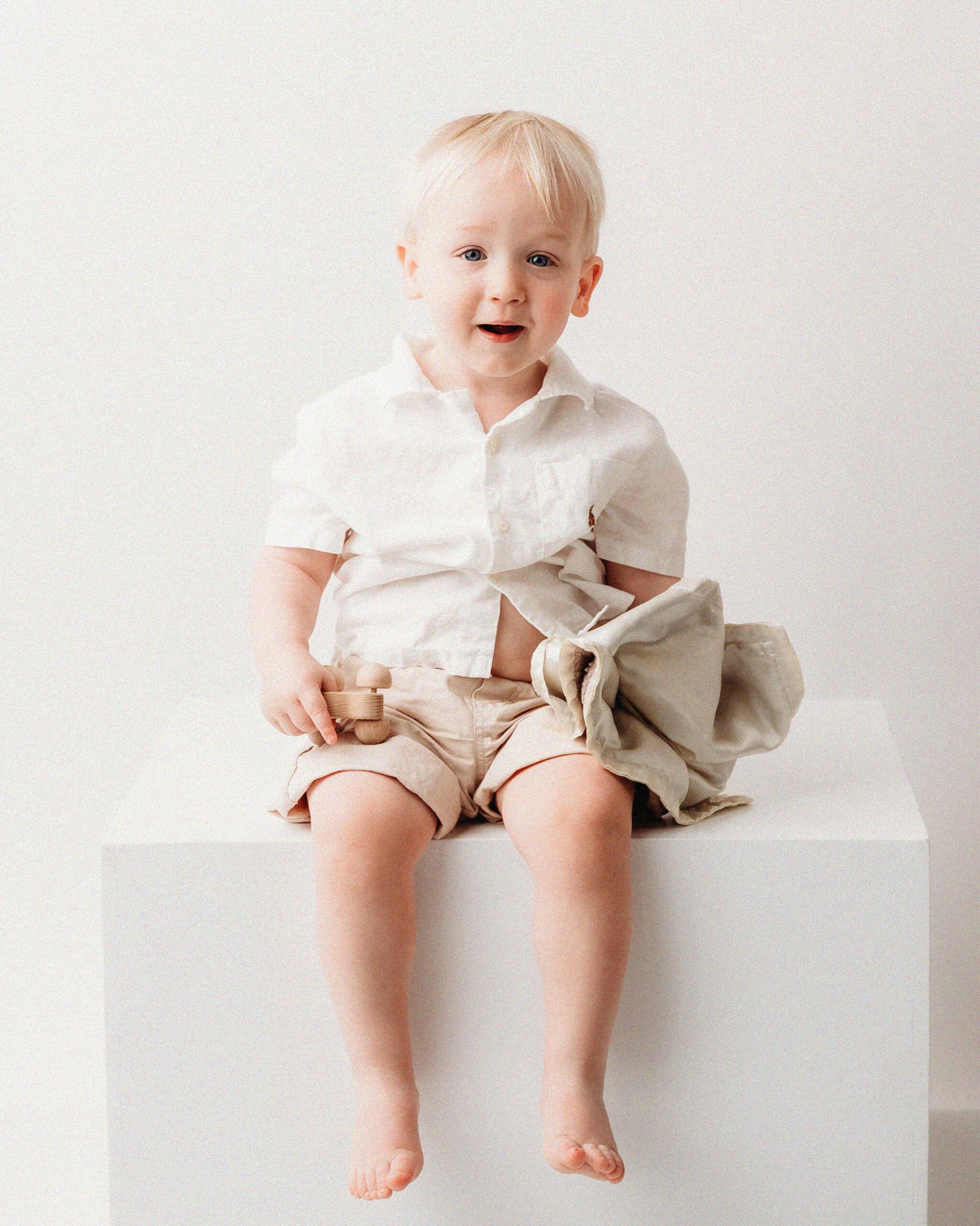 HILL_17 WEB Toddler boy in white shirt and khaki shorts sitting on a white cube during birthday portrait session at Kelli Dease Photography, Farmington CT