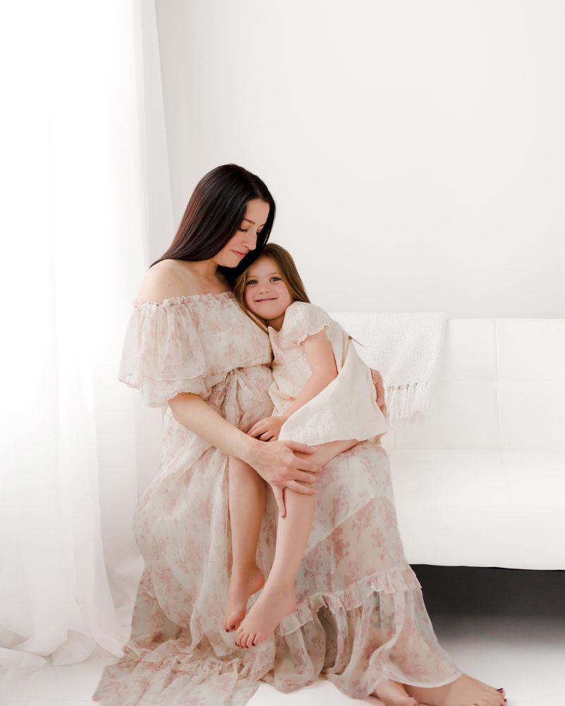 Pregnant mother in a blush off-shoulder gown holding her young daughter on her lap during a Farmington, CT maternity session with Kelli Dease Photography