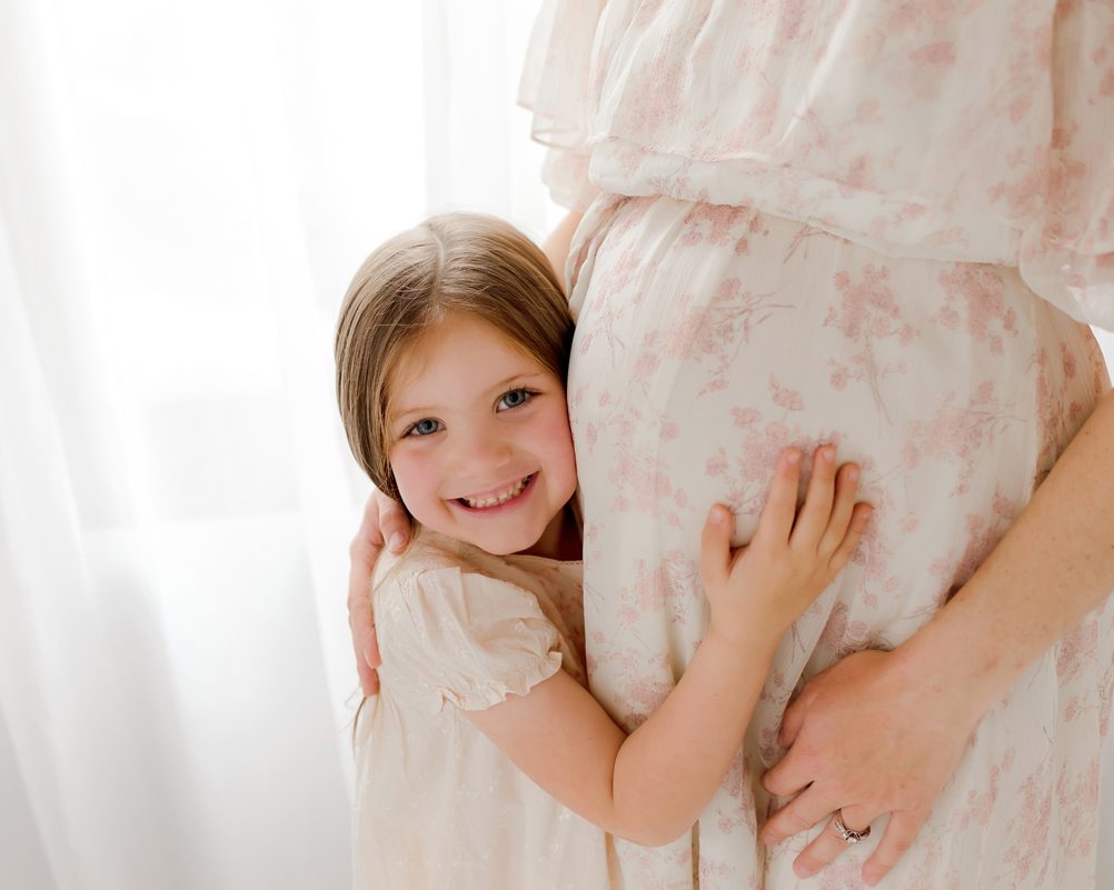 Young girl smiling as she hugs her mom's pregnant belly, one hand resting on the soft pink floral fabric