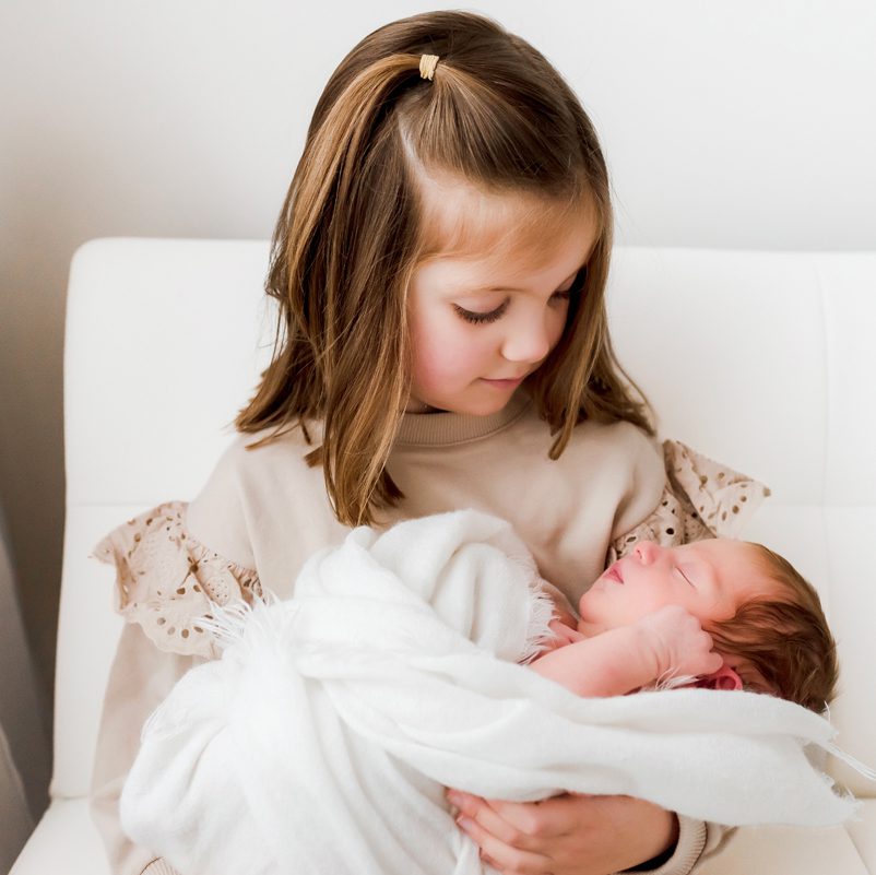 Big sister in a cream eyelet top cradling her swaddled newborn sibling on a soft white chair