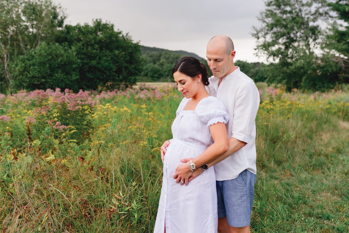 Pregnant woman and her partner standing together among pink wildflowers in a field, both looking down at her belly, taken during an outdoor maternity session in Simsbury, CT