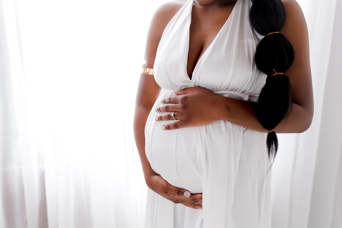 Close-up of a pregnant belly in a white deep-V gown, hands cradling the bump, soft window light during a studio maternity session at Kelli Dease Photography, Connecticut