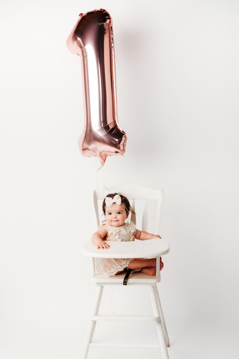 Baby girl sitting in a white high chair next to a rose gold number one balloon during a first birthday studio session at Kelli Dease Photography