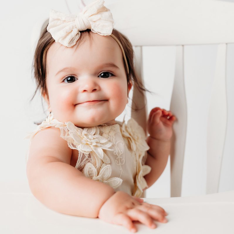 One-year-old girl in a cream lace romper smiling during her first birthday cake smash session at Kelli Dease Photography in Connecticut