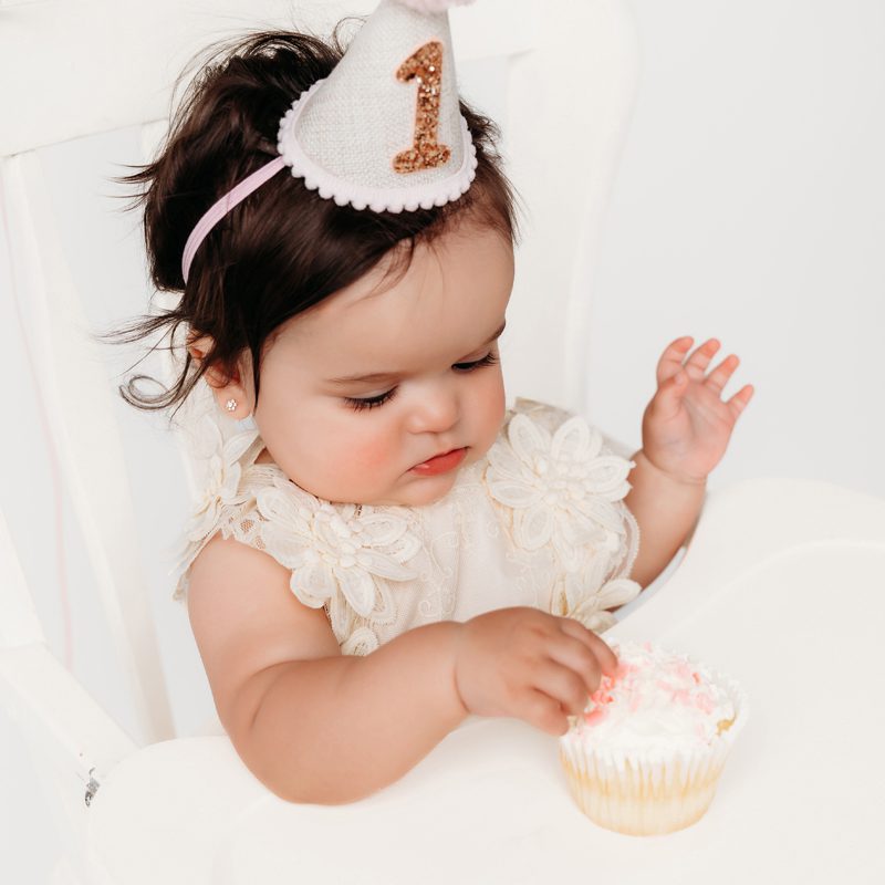 Baby girl in a white lace outfit reaching into her smash cake during a first birthday milestone session at Kelli Dease Photography