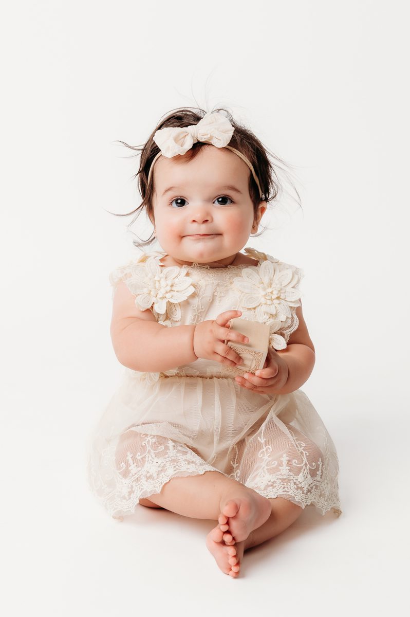 One-year-old girl in a cream lace dress sitting and smiling during her first birthday milestone session at Kelli Dease Photography
