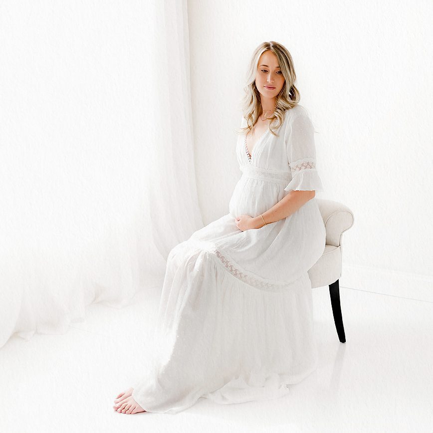 2024 Maternity Portfolio CT Photographer 1200px_COVER A pregnant woman sitting on a chair, holding her belly, in a white room during a maternity photo session at a Farmington, Connecticut photo studio.