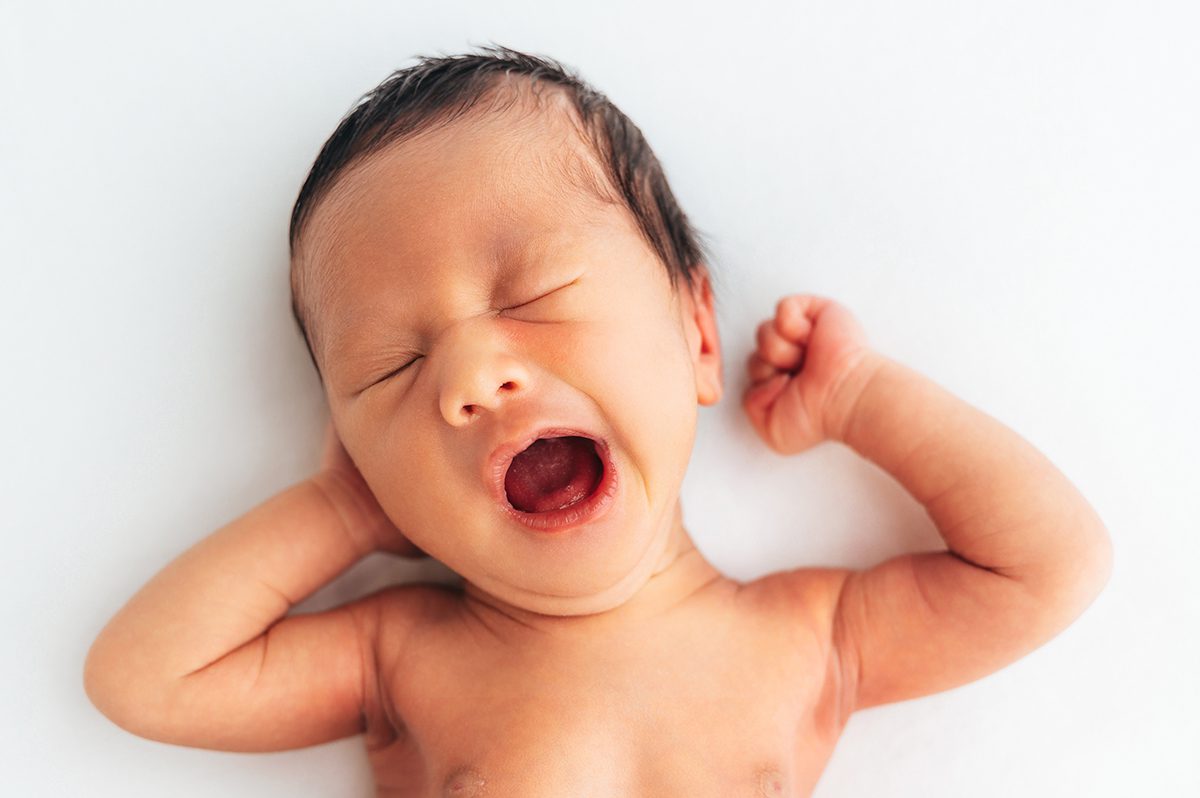Newborn baby mid-yawn on their back with one little fist raised, dark hair against a clean white backdrop, photographed during a studio newborn session with Connecticut newborn photographer Kelli Dease