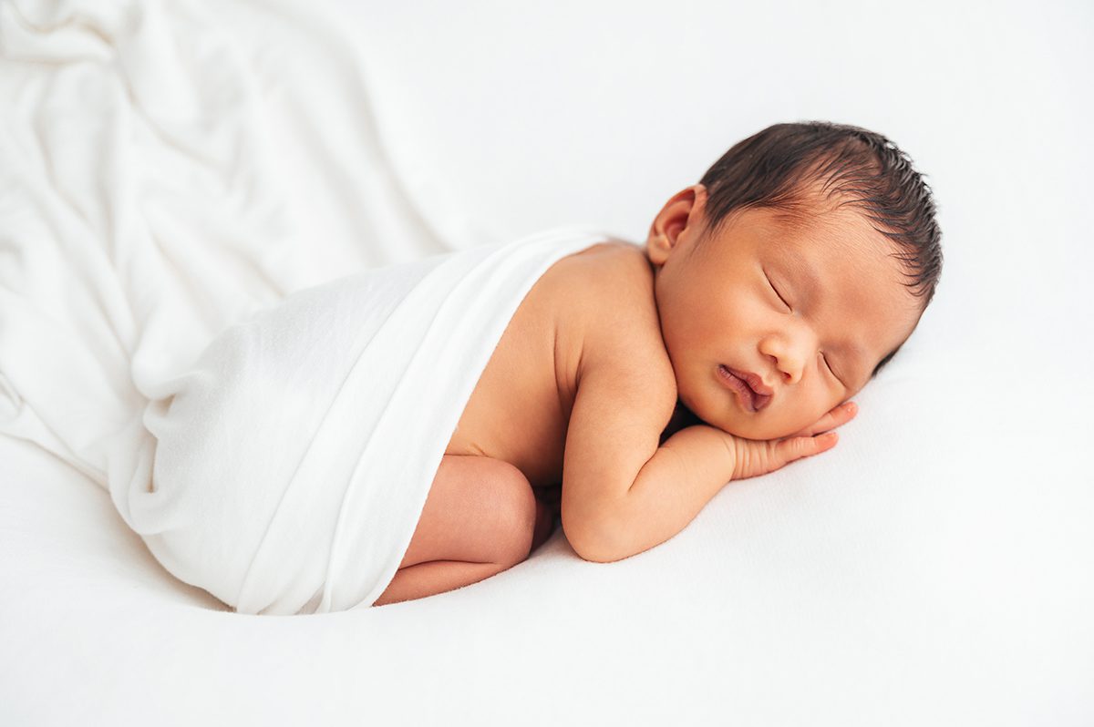 Newborn baby swaddled in a white wrap, sleeping peacefully curled on their side during a minimalist studio newborn session at Kelli Dease Photography in Farmington, CT