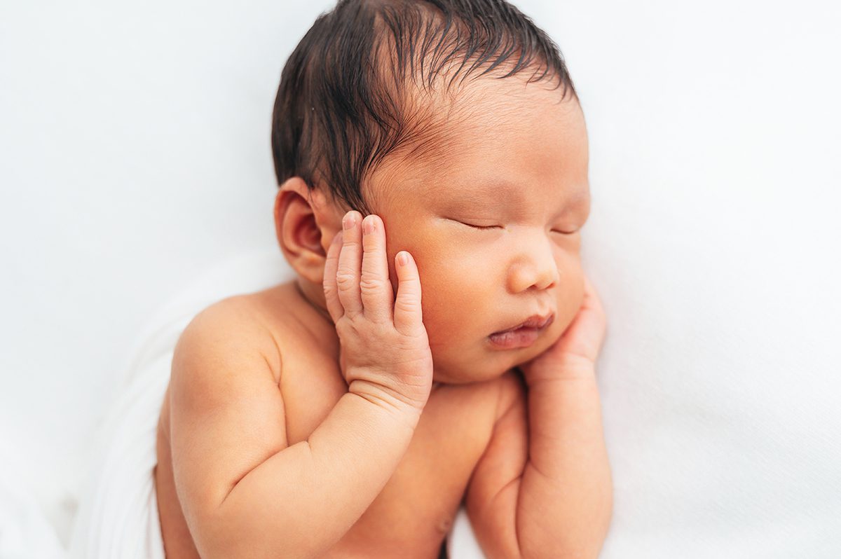 Sleeping newborn resting on their side with one hand tucked against their cheek, wrapped in soft white on a clean white backdrop, photographed by Farmington, CT newborn photographer Kelli Dease