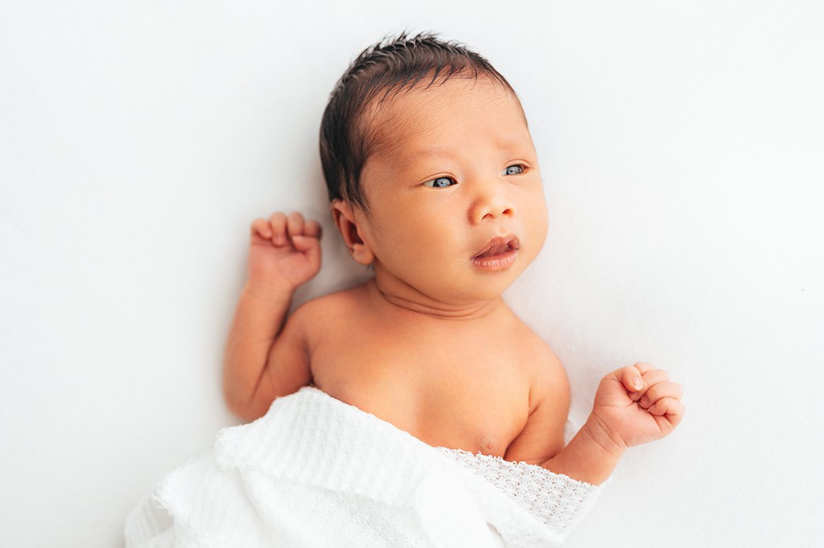 Wide-awake newborn lying on their back with bright eyes and arms stretched out, loosely wrapped in white knit fabric, photographed by Kelli Dease in Farmington, Connecticut