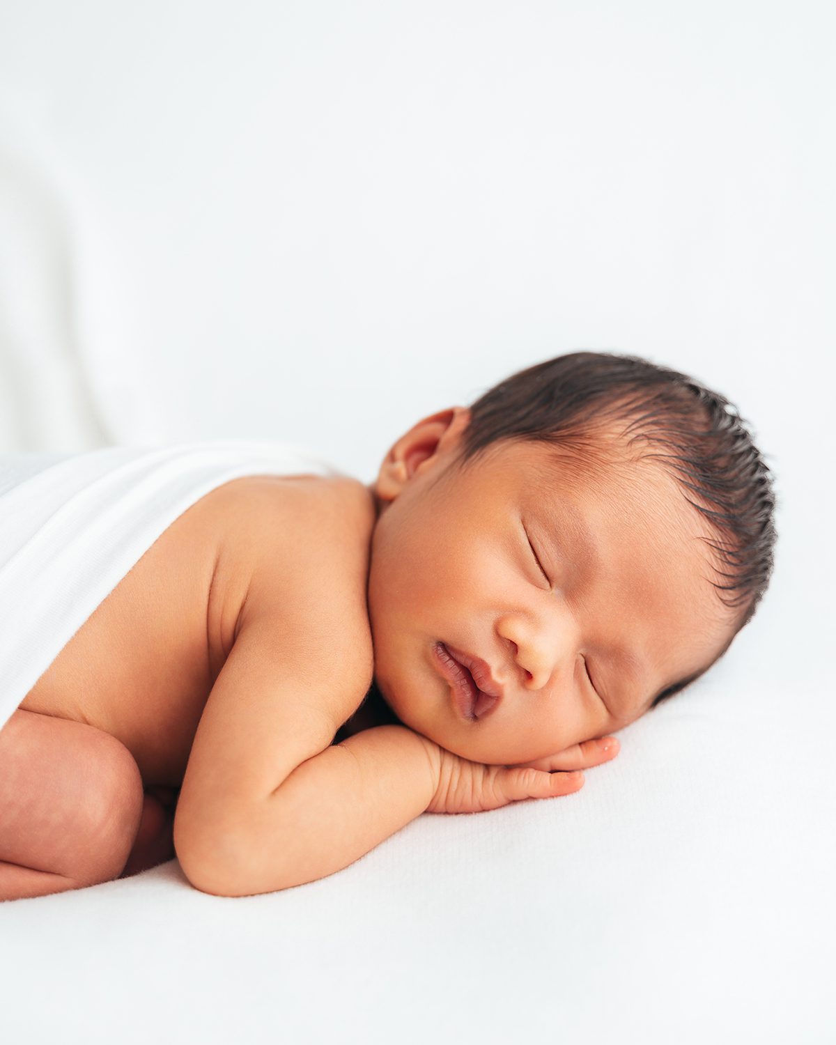 Sleeping newborn resting on their side with one hand tucked against their cheek, photographed against a clean white backdrop by Farmington, CT newborn photographer Kelli Dease