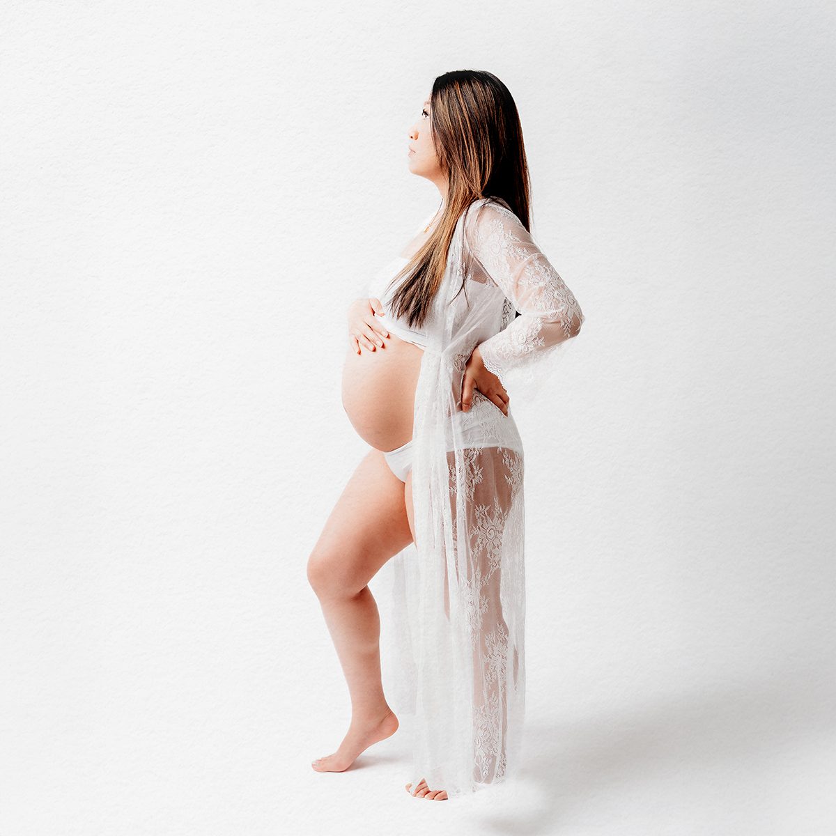 Farmington CT Studio Maternity Photography 03 Expectant mother in a white lace gown standing in profile against a clean studio backdrop, hand on her hip, at Kelli Dease Photography in Farmington, CT