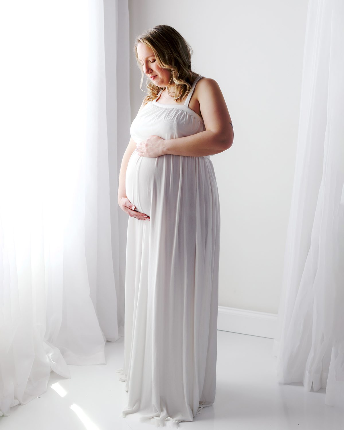 Farmington CT Studio Maternity Photography 05 Expectant mother in a white gown standing by a sunlit window with sheer curtains, hand resting on her bump, during a studio maternity session in Farmington, CT