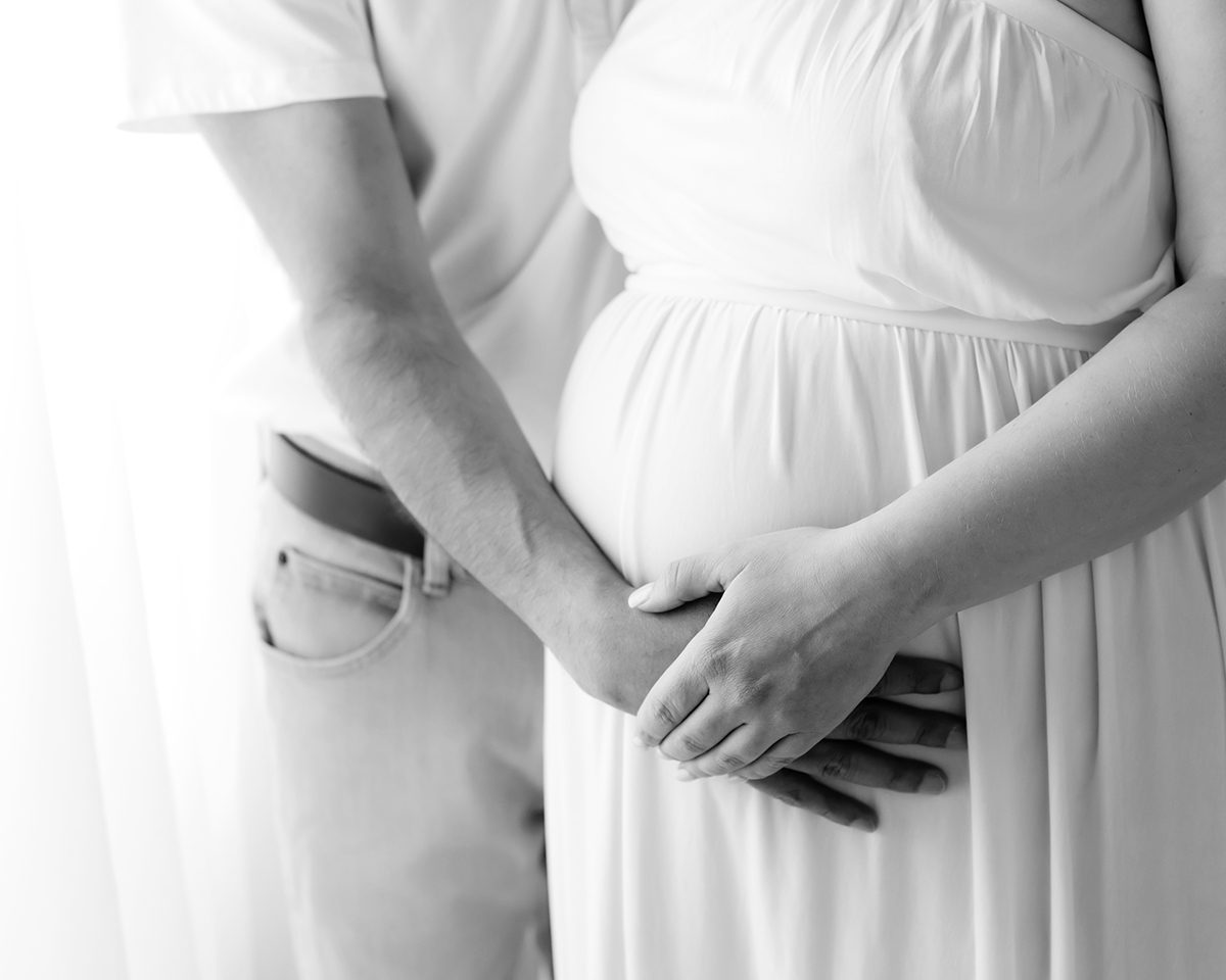 Farmington CT Studio Maternity Photography 06 Black and white close-up of an expectant couple's hands cradling the pregnant belly, mom in a white gown, at Kelli Dease Photography in Farmington, CT