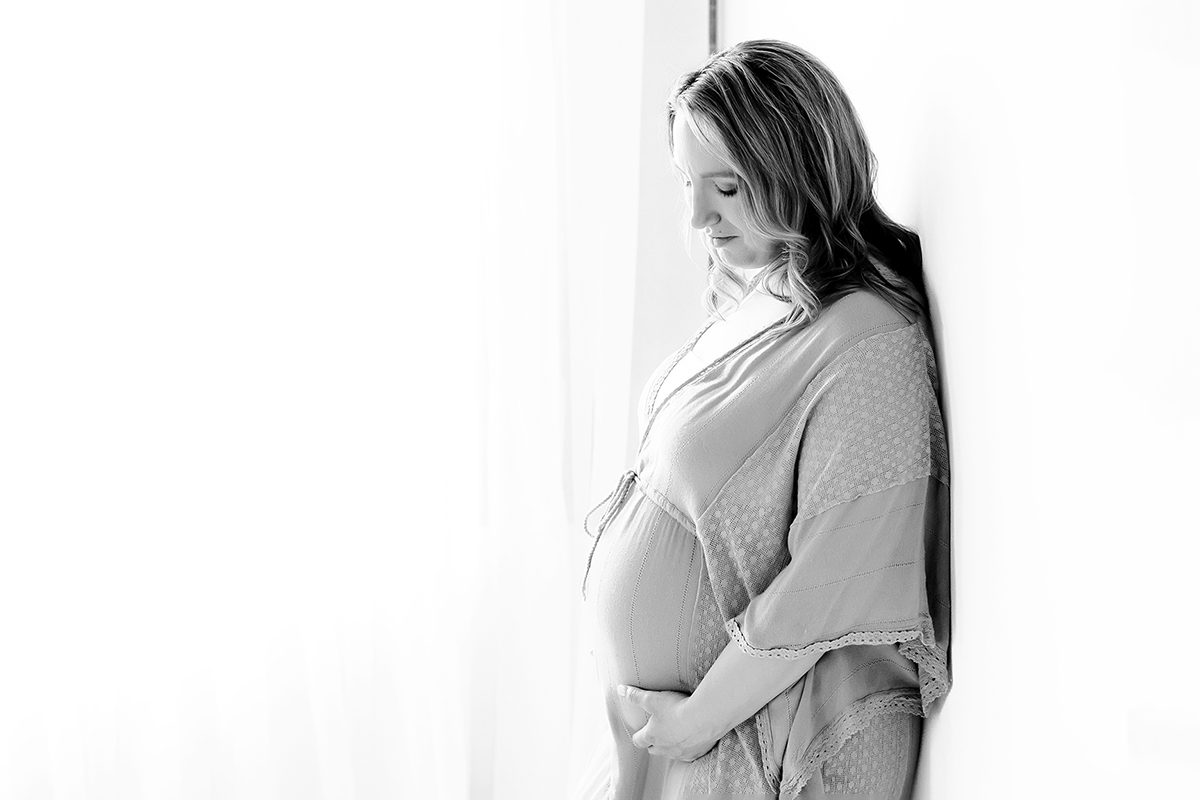 Black and white image of a pregnant woman standing next to a sheer colored window, cradling her belly in Kelli Dease's Farmington Connecticut Photography Studio