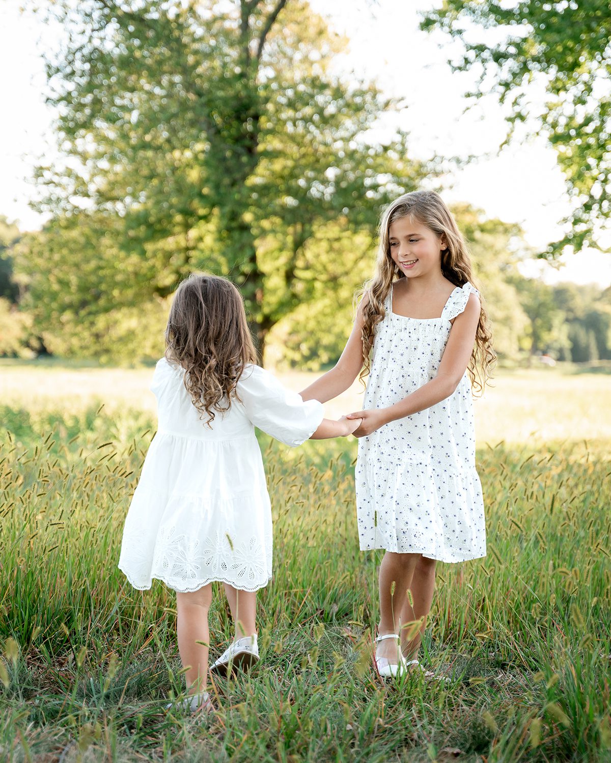 Two sisters in white summer dresses holding hands in a sunlit grassy field during an outdoor sibling session in Farmington, Connecticut